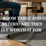 A person in jeans sitting on an office-style chair with casters at a wooden dining room table, positioned on a textured gray area rug. Overlaid text reads: "DINING ROOM TABLE AND CHAIRS WITH CASTERS: ARE THEY ACTUALLY WORTH IT FOR RENTERS?"