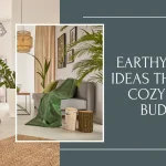 A blog post banner graphic featuring modern organic living spaces styled as an earthy room with a gray sofa, green throw blankets, wooden tree stump stools, potted indoor plants, and text reading "EARTHY ROOM IDEAS THAT FEEL COZY ON A BUDGET".