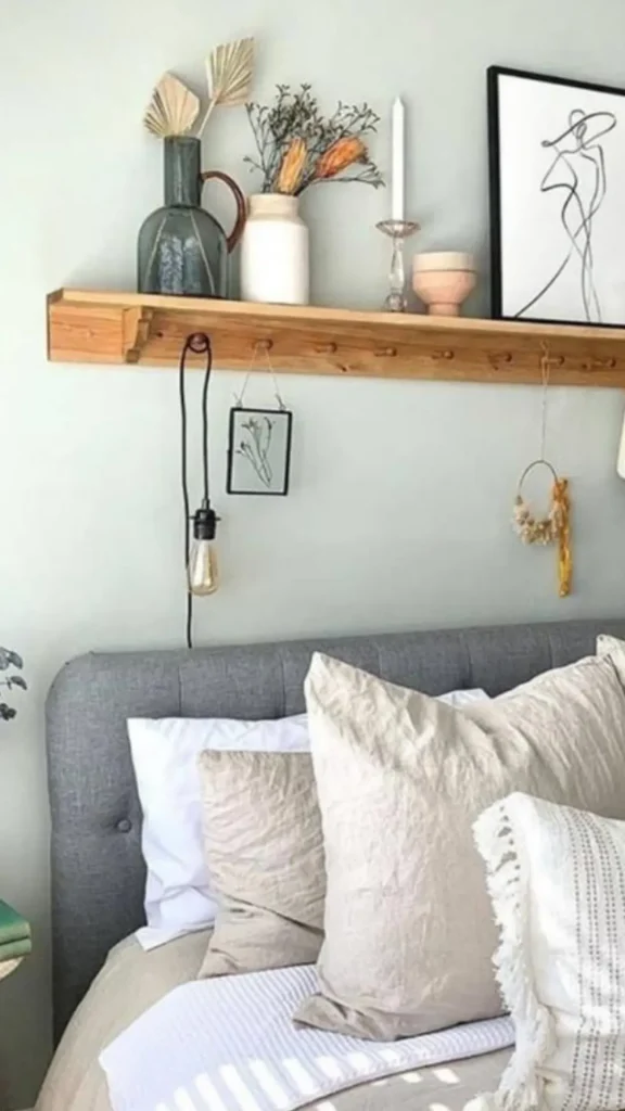 Smart small bedroom ideas utilizing a long wooden floating shelf with hanging pegs directly above a tufted grey headboard.